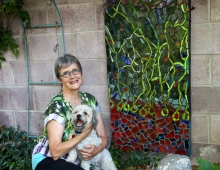 A woman holding a small dog, kneeling in a garden next to a colorful mosaic.